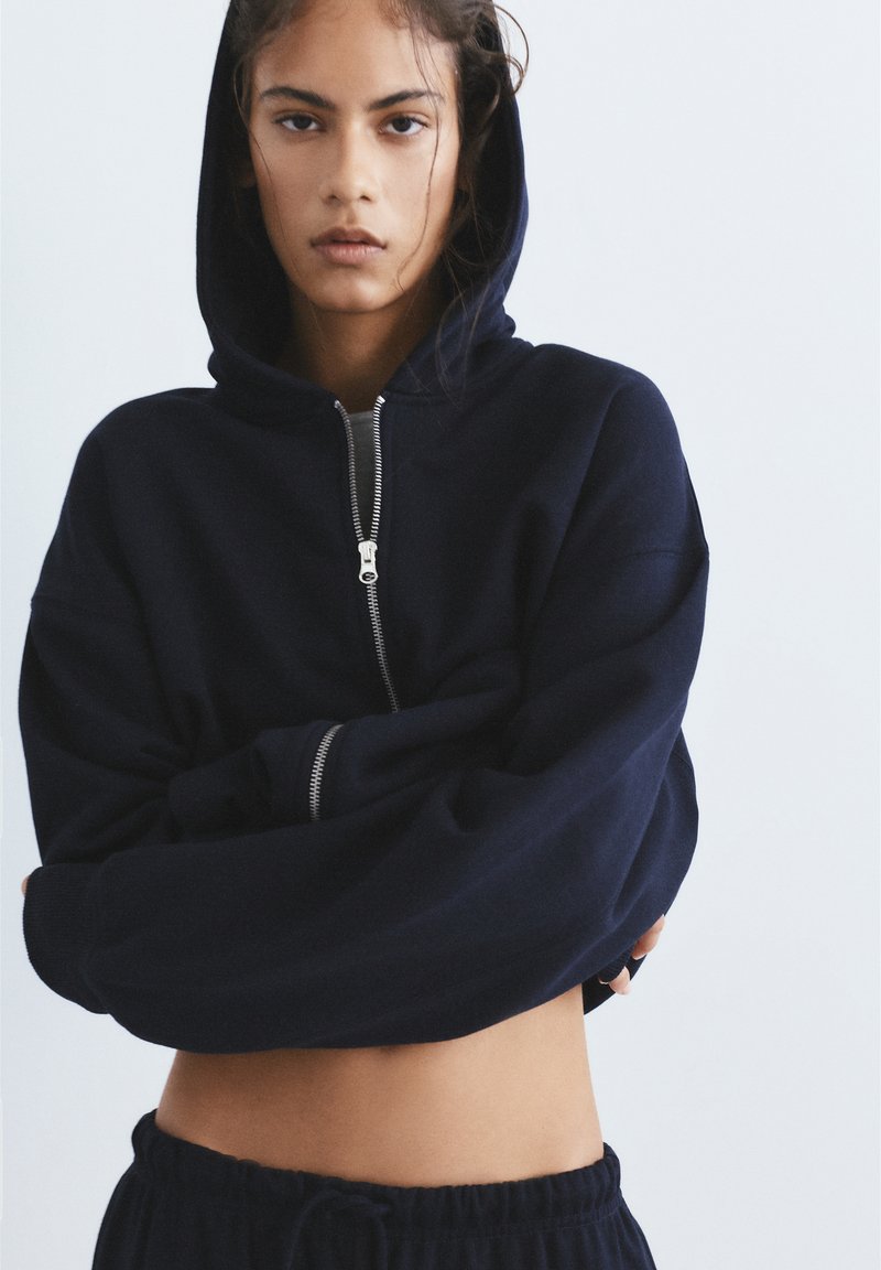 Tânăr cu păr închis la culoare, purtând un hanorac albastru închis scurt cu fermoar și glugă ridicată, cu brațele încrucișate și pantaloni negri cu șnur, pe un fundal simplu.