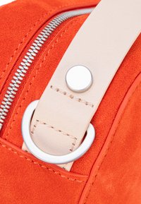 Close-up van een oranje suède tas met een zilveren rits, een beige band met een cirkelvormig zilveren detail en stikselaccenten.