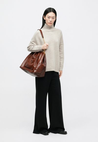 Γυναίκα που φοράει μπεζ πουλόβερ με ζιβάγκο και μαύρο παντελόνι φαρδιάς γραμμής, κρατώντας μεγάλο καφέ δερμάτινο τσάντα ώμου μπροστά σε λευκό φόντο.