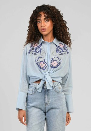 Femme aux cheveux bouclés portant une chemise bleue claire boutonnée et nouée à la taille avec une broderie florale et un jean taille haute bleu clair.