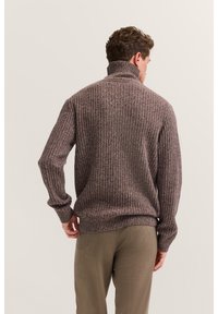 Brauner gerippter Pullover mit hohem Kragen und langen Ärmeln, der über texturreiche Variationen verfügt. Wird über hellen, enganliegenden Hosen getragen.