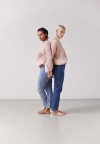 Två personer står rygg mot rygg, iklädda överdimensionerade räfflade rosa tröjor och blårandiga jeans, barfota på en neutral bakgrund.