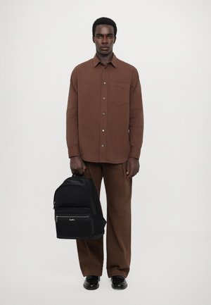 Camicia marrone con bottoni e taschino sul petto, abbinata a pantaloni marroni. Tien un zaino nero con cerniera argentata e dettagli con logo.