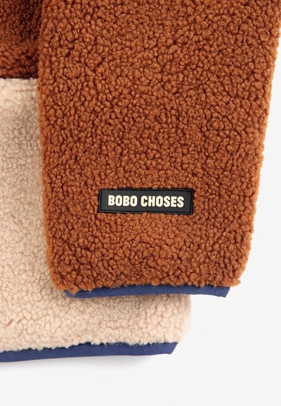 Deux manches en tissu polaire, une marron et une beige, toutes deux avec des ourlets bleu marine, la manche marron porte une étiquette noire avec l'inscription BOBO CHOSES.