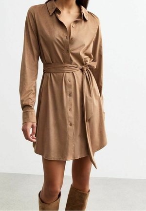 Femme portant une robe-chemise marron clair à manches longues avec des boutons sur le devant, une ceinture nouée à la taille et des bottes assorties montant jusqu'aux genoux.