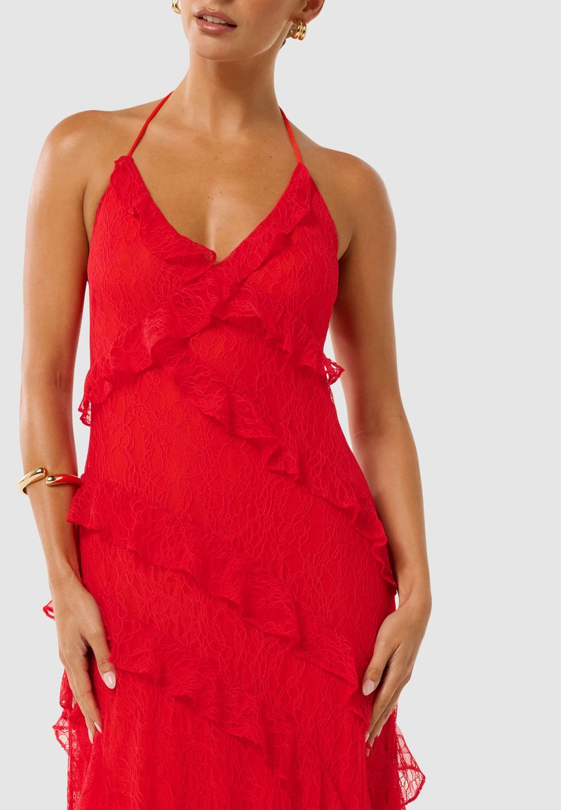 Robe en dentelle rouge avec un décolleté en V profond, ornée de volants superposés et de bretelles spaghetti. Texture lisse avec une silhouette fluide. Accent bracelet en or.