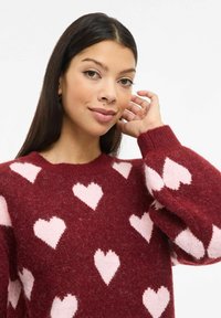 Crveni džemper s ružičastim uzorcima srca, okruglim izrezom i rebrastim manšetama. Mekana tekstura, srednje težine, ležerni kroj.