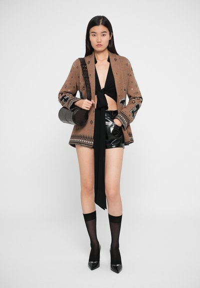 Gilet marron brodé avec des motifs géométriques, top court noir, shorts noirs brillants, chaussettes noires transparentes et escarpins noirs pointus.