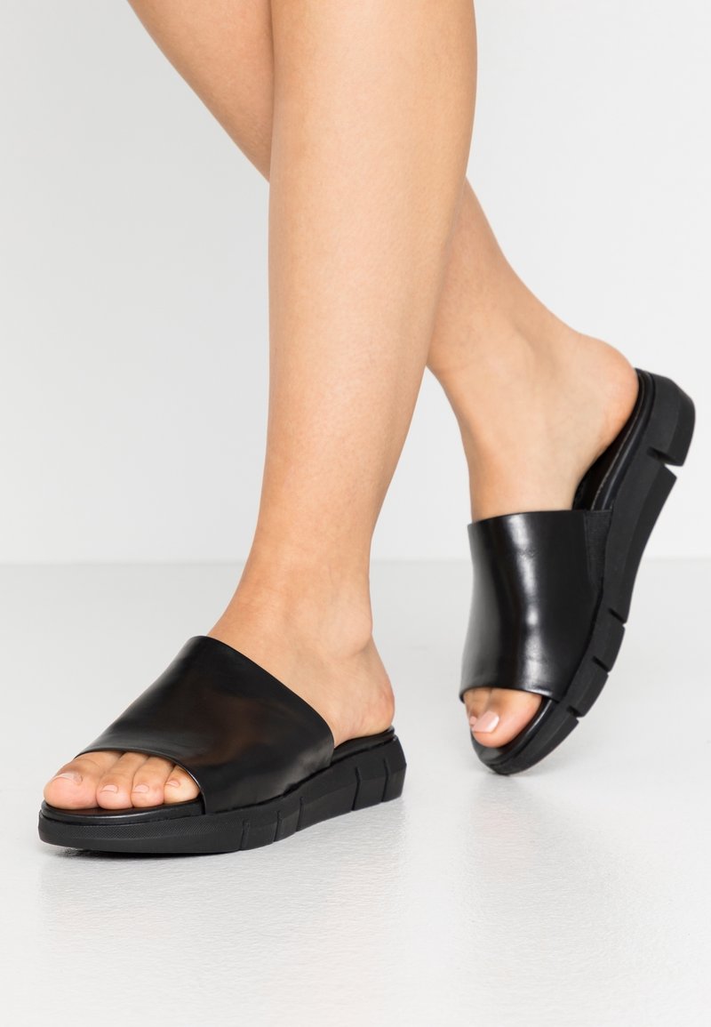 Les sandales à glissière noires présentent une tige en cuir lisse et brillant ainsi qu'une semelle en caoutchouc épaisse et texturée avec un design minimaliste. Style à bout ouvert.