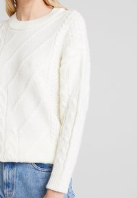 Femme portant un pull en maille torsadée blanc avec des motifs en losange texturés et un jean bleu clair, sur un fond clair uni.