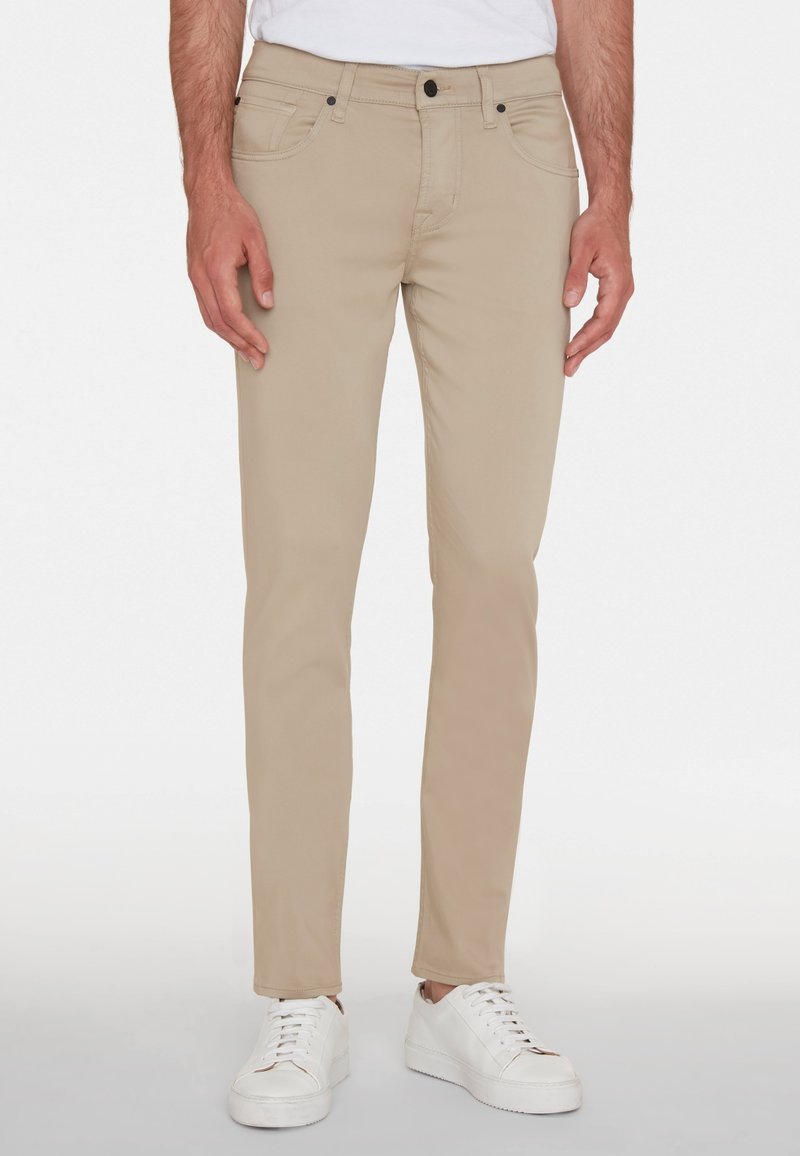Beige Slim-Fit-Jeans aus einem Mischgewebe, ausgestattet mit vorderen und hinteren Taschen, Kontrastnähten und einem Knopfverschluss. Tragen mit weißen Sneakern.