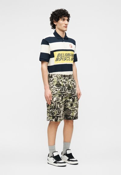 Νεαρός άνδρας όρθιος, φοράει μαύρο-άσπρο ριγέ πόλο με κίτρινο κείμενο "Billionaire Boys Club", παραλλαγής cargo σορτς, γκρι κάλτσες και μαύρα-άσπρα αθλητικά παπούτσια.