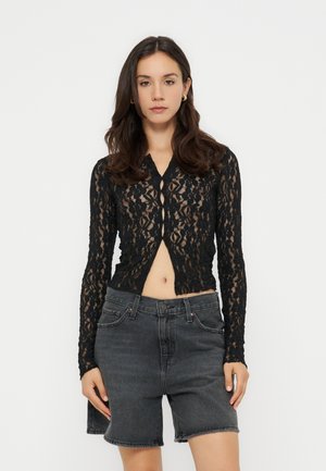 Frau mit langen dunklen Haaren, die ein schwarzes Spitze-Langarmshirt und hoch taillierte dunkelgraue Jeansshorts trägt und vor einem weißen Hintergrund steht.