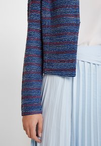 Teksturowana, krótka kurtka w niebieskie, czerwone i białe paski, zestawiona z jasnobłękitną plisowaną spódnicą. Gładka tkanina kontrastuje z prążkowaną powierzchnią.