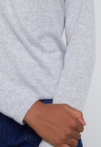 Suéter de punto gris claro con textura y mangas largas, con un ajuste ceñido en los puños, llevado sobre mezclilla oscura.