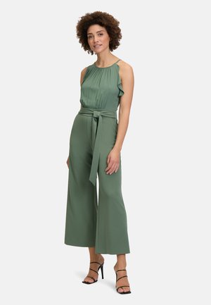 Grüner Jumpsuit mit Rüschensaum am Hals, tailliertem Bund und weitem Bein. Glatte Textur mit fließendem Design, ergänzt durch Riemchen Sandalen.