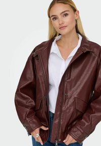 Giacca in pelle bordeaux con vestibilità rilassata, chiusura con zip, bottoni a pressione e tasche frontali. Texture liscia e design oversize.