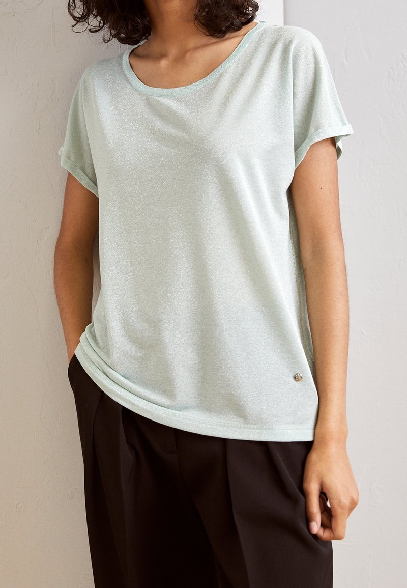 Femme portant un haut vert menthe clair à manches courtes légèrement rentré dans un pantalon noir plissé, debout devant un mur texturé.