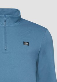 Blauer Pullover mit Reißverschluss bis zur Brust und hohem Kragen sowie kleinem rechteckigem schwarzen Logoaufnäher auf der linken Brustseite.
