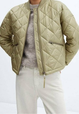Beige quiltet bomberjakke med lynlåslommer båret over en lysegrå sweater og off-white bukser.