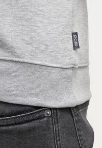 Primer plano del dobladillo de un sudadera gris con la etiqueta KOM encima del bolsillo de unos jeans de mezclilla negros y un botón de metal.