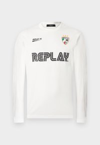 Hvid langærmet skjorte med sort "REPLAY" tekst henover brystet, lille "REPLAY FC" tekst øverst til venstre, og et tri-farvet skjold emblem med tre stjerner øverst til højre.