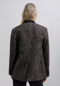 Blazer de tweed con patrón de pata de gallo en tonos marrón y gris, que presenta una abertura en la parte trasera y un cuello negro, con mangas largas y un corte entallado.