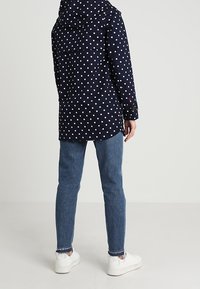 Marinblå prickig kappa med huva, något längre modell, kombinerad med blå jeans och vita sneakers. Kappan har dragkedja och fickor.