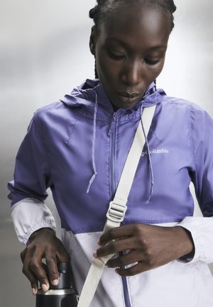 Persoon in paarse en witte jas die een witte band verstelt terwijl hij een zwarte thermosfles vasthoudt.