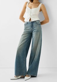 Bílý bavlněný cropped top s širokými ramínky a předními knoflíky, spárovaný se světle modrými širokými džínami s vybledlými místy. Špičaté bílé podpatky.