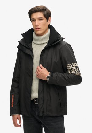 Veste imperméable noire avec un col haut, arborant le logo "Superdry" beige sur la manche et un accent orange sur le poignet. Portée par-dessus un col roulé gris.