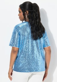 Top corto a maniche corte in blu chiaro con paillettes, caratterizzato da una superficie lucida e una vestibilità ampia. La vista posteriore mette in risalto la texture brillante e l'orlo arrotondato.