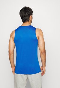 Hombre vestido con una camiseta atlética sin mangas de color azul brillante y pantalones de chándal gris claro, de pie con la espalda hacia la cámara.