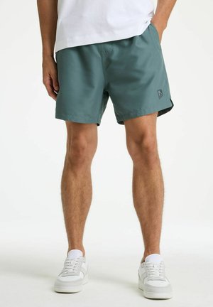 Mees, kandes türkiissiniseid lühikesi pükse ja valgeid tossusid, seisab ühe käega taskus ühevärvilise tausta ees.