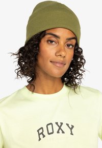Gorro de canalé verde oliva con un ajuste ceñido, combinado con una camiseta amarilla clara que presenta un logo impreso oscuro. Cabello rizado visible.