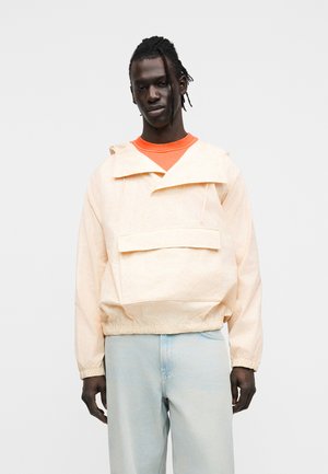 Light peach hooded pullover jacket with front pocket, elastic cuffs, and hem; worn over an orange shirt, paired with light blue jeans.