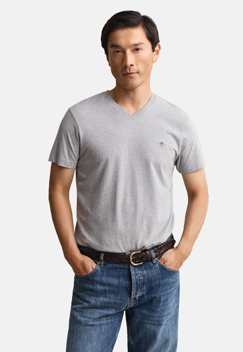 Hombre con camiseta de cuello en V gris claro, vaqueros azules y cinturón trenzado marrón, de pie con las manos en los bolsillos frente a un fondo liso.