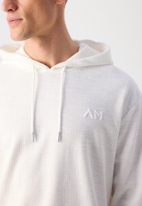 Hoodie bianco con trama, dotato di tasca frontale, cappuccio con cordoncino e un piccolo logo tono su tono. Il tessuto presenta un motivo delicato e una finitura morbida.