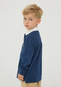 Granatowa koszula z długim rękawem z białym kołnierzykiem i bocznymi rozporkami. Miękka faktura z detalem logo na piersi. Swobodny krój dla dzieci.