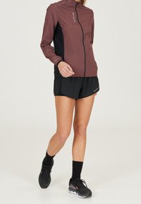 Bordowa i czarna kurtka biegowa z wysokim kołnierzem, pełnym zamkiem błyskawicznym oraz odblaskowymi elementami, w połączeniu z czarnymi sportowymi szortami i butami sportowymi.