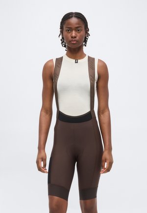 Žena v bielom tielku bez rukávov a hnedých cyklistických kraťasoch s trakmi stojí pred jednoliatym bielym pozadím.