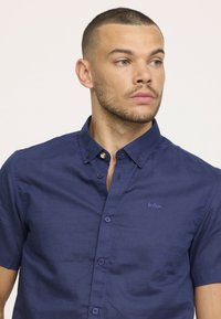 Chemise à manches courtes bleu marine en tissu texturé, dotée d'un col boutonné, de boutons sur le devant et d'un logo brodé sur la poitrine.