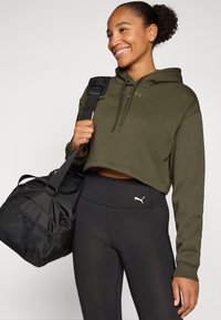 Camisola curta verde-oliva com cordões, combinada com leggings pretas de cintura alta com um logo branco da Puma, e uma mala de ginásio preta.