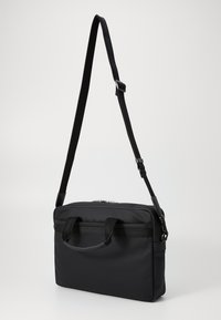 Sac noir en matériau synthétique, de forme rectangulaire, avec des doubles poignées supérieures et une bandoulière réglable. Présente une fermeture éclair.