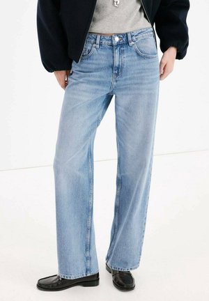Persoon die lichtblauwe jeans met rechte pijpen, zwarte schoenen, een grijs top en een zwart jasje draagt, staand op een witte vloer.