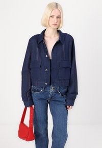 Ναυτική cropped ζακέτα με δύο μπροστινές τσέπες, ορατές ραφές σε κόκκινο χρώμα, συνδυασμένη με μπλε τζιν και μικρή κόκκινη τσάντα.