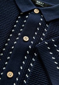 Polo en tricot navy avec un tissu texturé, des boutons en bois et des coutures décoratives blanches le long de la patte de boutonnage et du col.