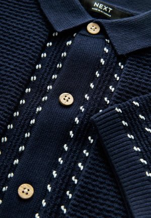 Polo en tricot navy avec un tissu texturé, des boutons en bois et des coutures décoratives blanches le long de la patte de boutonnage et du col.