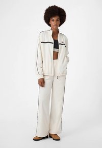 Creme kleurig trainingsjack met ritssluiting en zwarte strepen, gecombineerd met bijpassende losse broek. De outfit bevat een cropped top eronder voor laagjes.