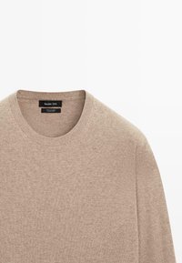 Beige kashmirtröja med rund halsringning. Mjuk textur, minimalistisk design och subtilt fläckmönster, med en svart varumärkesetikett.